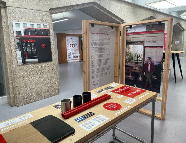 A wooden exhibition table with materials and captions, in front of a timber frame with information panels and an image of the new TfL bus shelter design in The London Centre gallery
